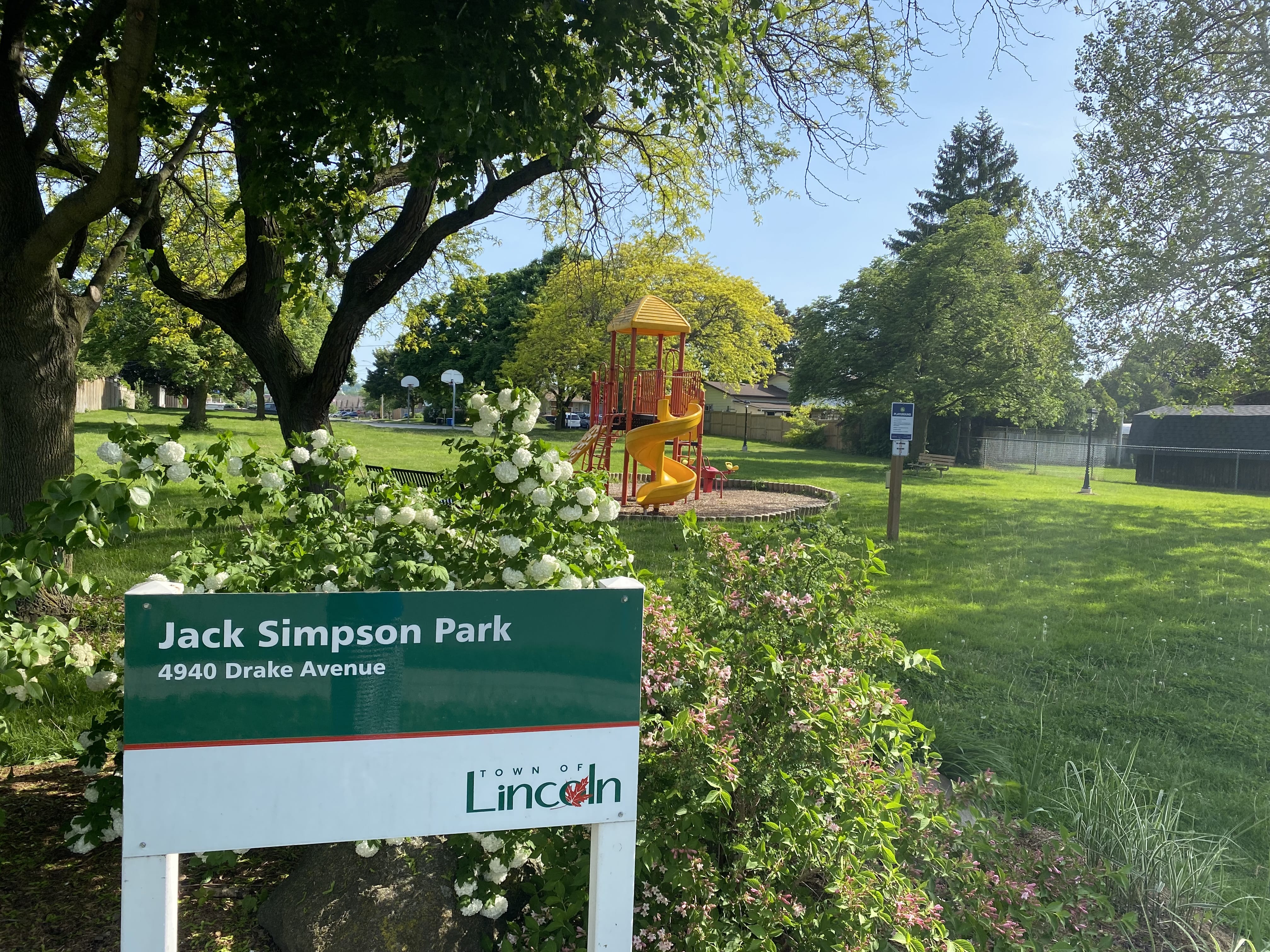 Sign with "Jack Simpson Park 4940 Drake Avenue" Written on it. The sign is situated near a park. There is a yellow spiral slide in the background.