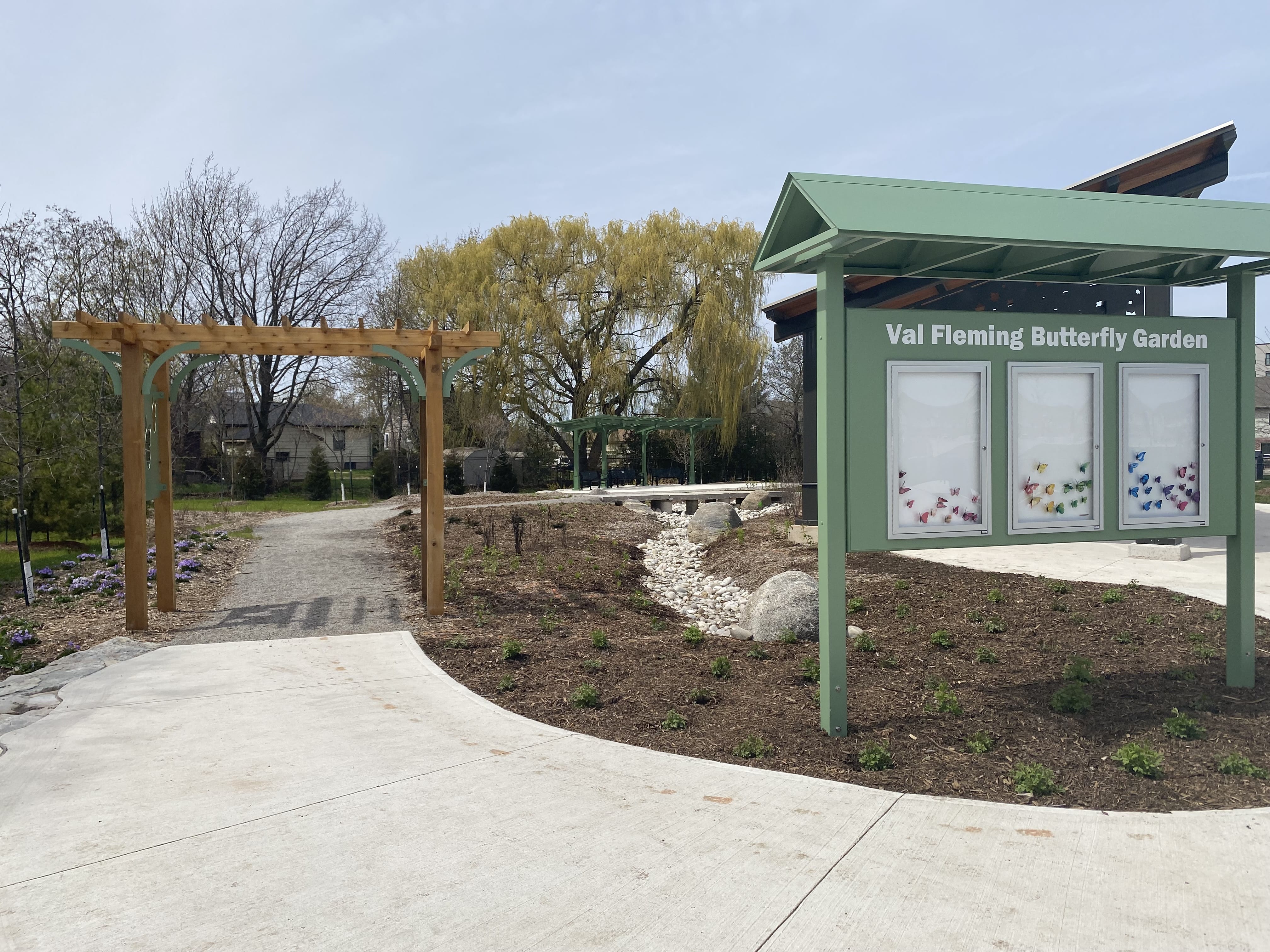 Community board that reads "Val Fleming Butterfly Garden." The community board is by the side of a concrete pathway.