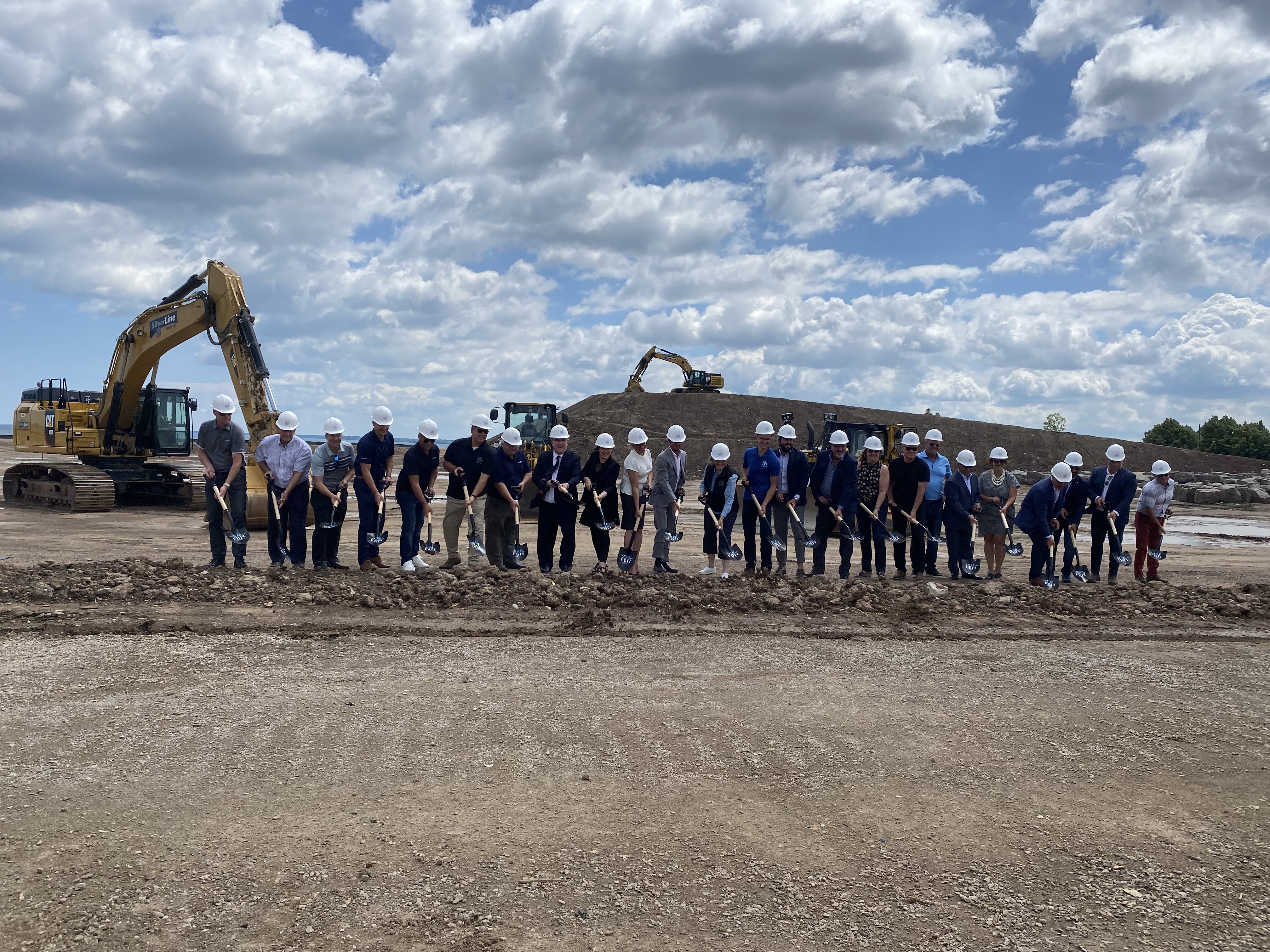 A line of about 25 adults standing on dirt wearing white hard hats pose with shovels as if they are about to dig.