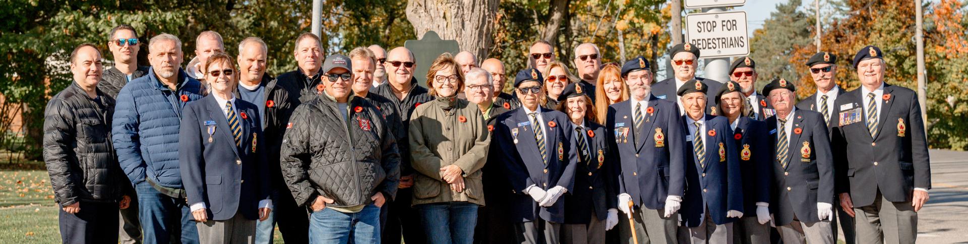 Group of people and veterans in uniform standing on the edge of the sidewalk in front of a painted crosswalk.