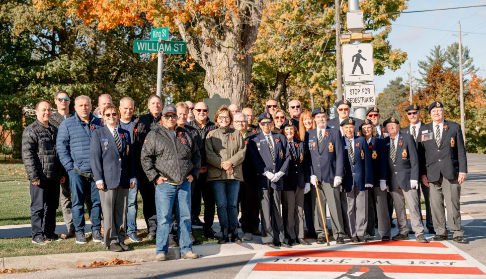 Group of people and veterans in uniform standing on the edge of the sidewalk in front of a painted crosswalk.