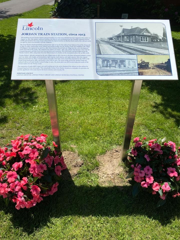 Plaque detailing the history of the Jordan Train Station. Two flower bushes flank the plaque's sides.