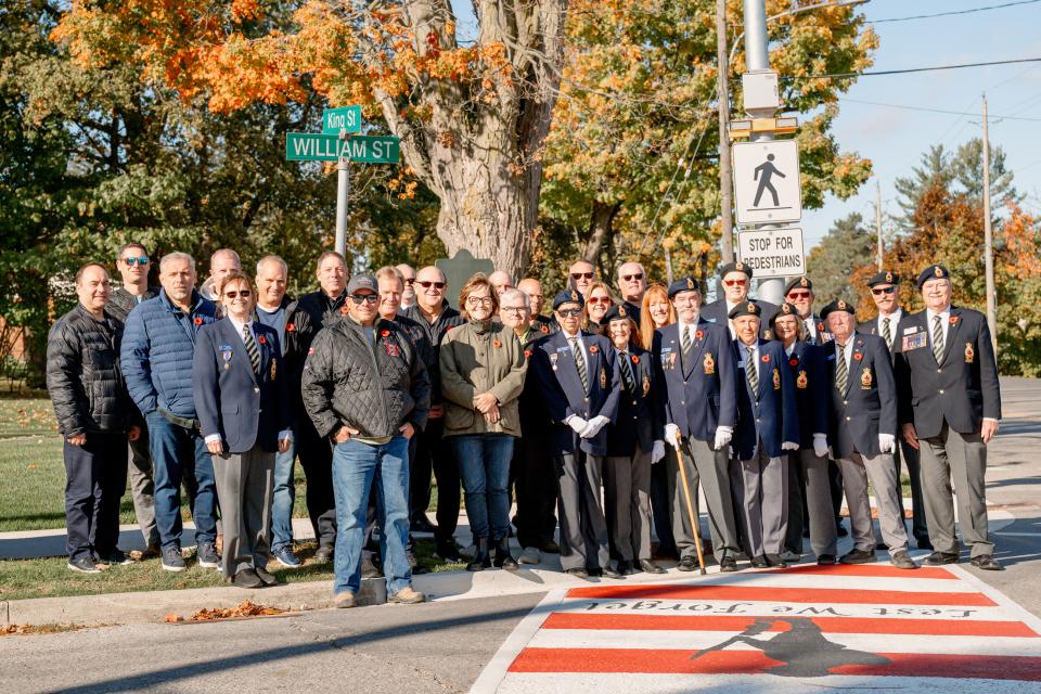 Group of people and veterans in uniform standing on the edge of the sidewalk in front of a painted crosswalk.