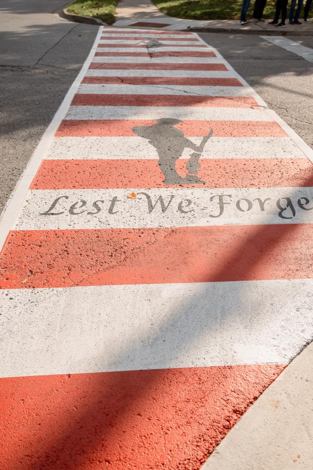 Crosswalk painted with red and white paint with a soldier's silhouette and words painted on crosswalk.