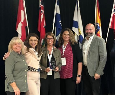 Five people in front of flags with middle person holding award